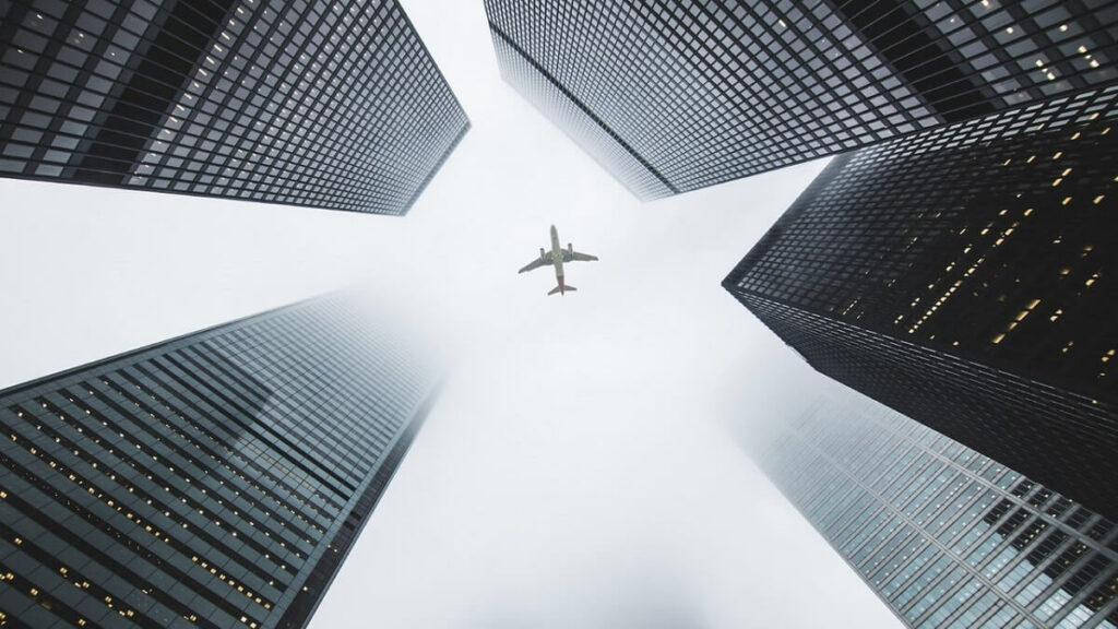 Imagem preta e branca de um avião sobrevoando edifícios altos, com nuvens cobrindo os andares superiores. Bug de passagens aéreas 2022