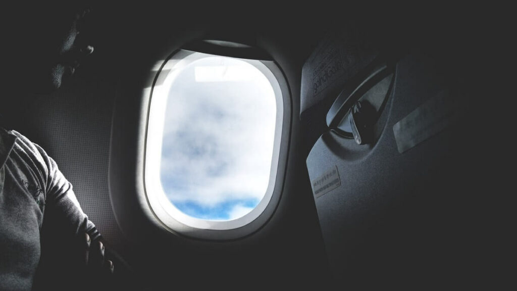 Imagem interna preta e branca de um homem olhando o céu azul pela janela de um avião